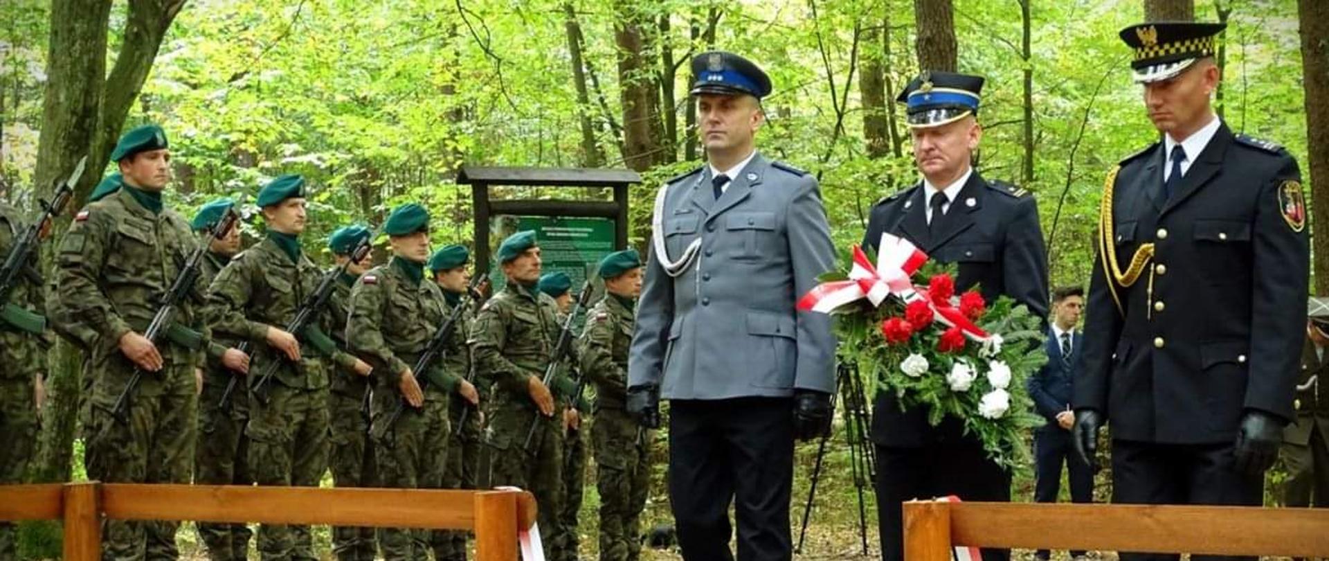 Zdjęcie przedstawia delegację służb mundurowych składającą kwiaty. Od lewej stoi policjant w umundurowaniu wyjściowym, w środku strażak mł. bryg. Łukasz Drzewiecki ubrany w mundur wyjściowy trzymający kwiaty, po prawej strażnik miejski. W tle widoczny pododdział żołnierzy Wojska Polskiego. 