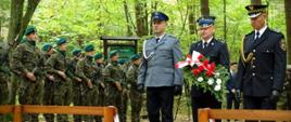 Zdjęcie przedstawia delegację służb mundurowych składającą kwiaty. Od lewej stoi policjant w umundurowaniu wyjściowym, w środku strażak mł. bryg. Łukasz Drzewiecki ubrany w mundur wyjściowy trzymający kwiaty, po prawej strażnik miejski. W tle widoczny pododdział żołnierzy Wojska Polskiego. 
