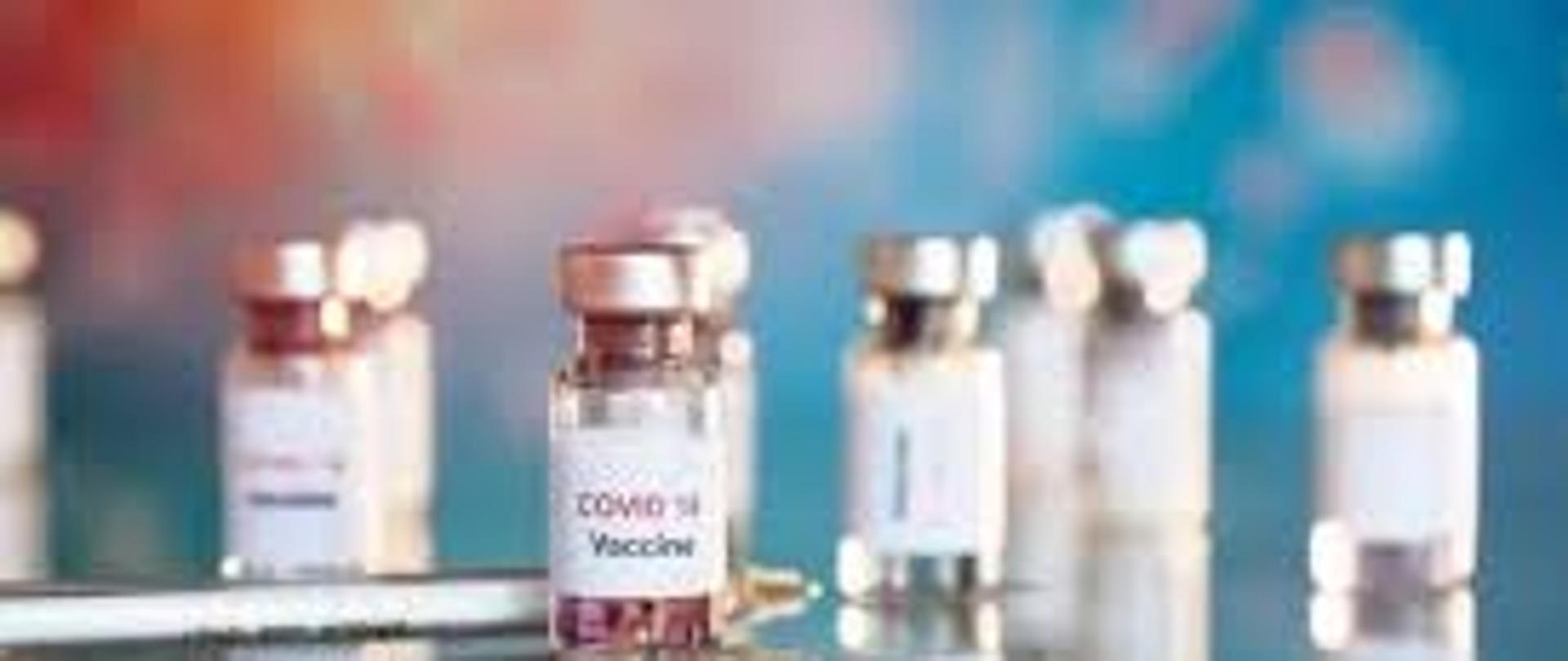 Zdjęcie przedstawia pojemniki ze szczepionką na covid-19 oraz strzykawkę umieszczone na stole.