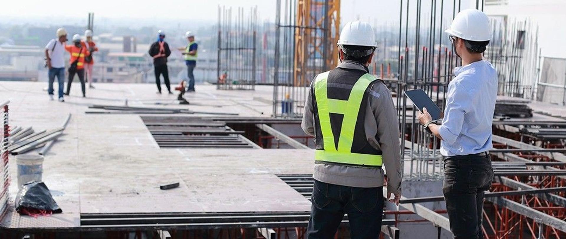 Dwie osoby w białych kaskach ochronnych rozmawiające na placu budowy podczas słonecznego, wiosennego dnia