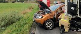 Widok na rozbite auto. Widać jego przód. Auto jest mocno uszkodzone. Zdjęcie wykonane dzień. Przy aucie stoi strażak.