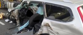 Wypadek na obwodnicy Rawicza. Na pierwszym planie srebrny samochód osobowy, bardzo mocno zniszczony (w szczególności przód i lewa strona). W tle widoczny jest fragment zestawu pojazdów: ciągnika siodłowego i naczepy. 