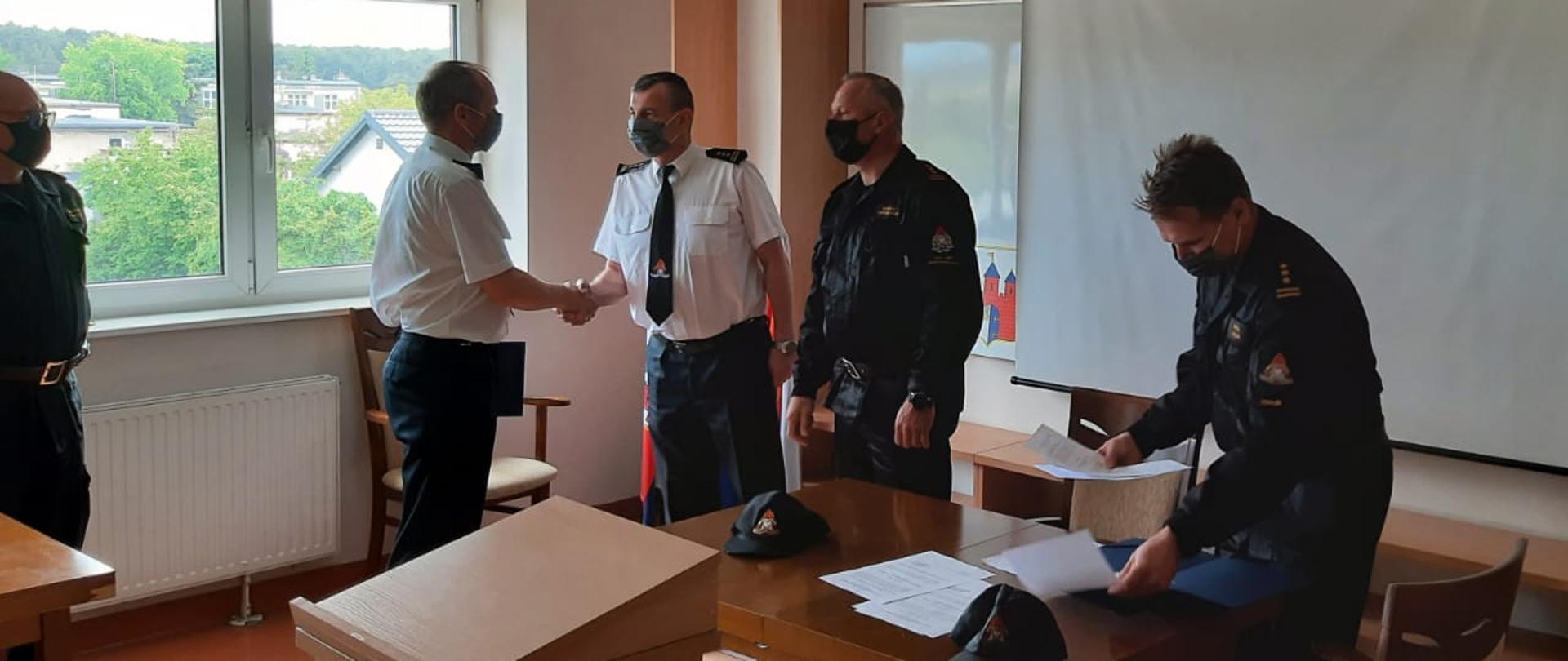 W centralnej części zdjęcia mł. bryg. Tomasz Czajka ściskający rękę st. bryg. Janisławowi Bullerowi, z prawej strony st. bryg. Robert Wiśniewski, st. bryg. Marek Rusoń, z lewej strony st. bryg. Jarosław Jakub Koprowski, wszyscy maseczki na twarzach, osoby znajdują się w świetlicy JRG2 Bydgoszcz