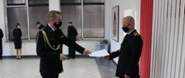 Dwóch strażaków - komendant wręczający dokument strażakowi emerytowi, na ścianie flaga państwowa i logo PSP