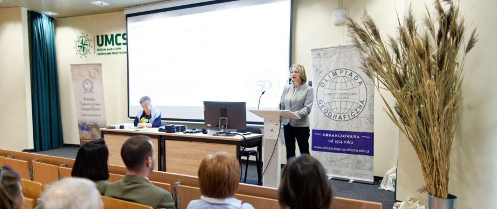 Aula UMCS. Widok na mównicę, przy której stoi kobieta. Za jej plecami, na ścianie rzutnik z prezentacją, którą oglądają siedzący na widowni uczestnicy olimpiady.