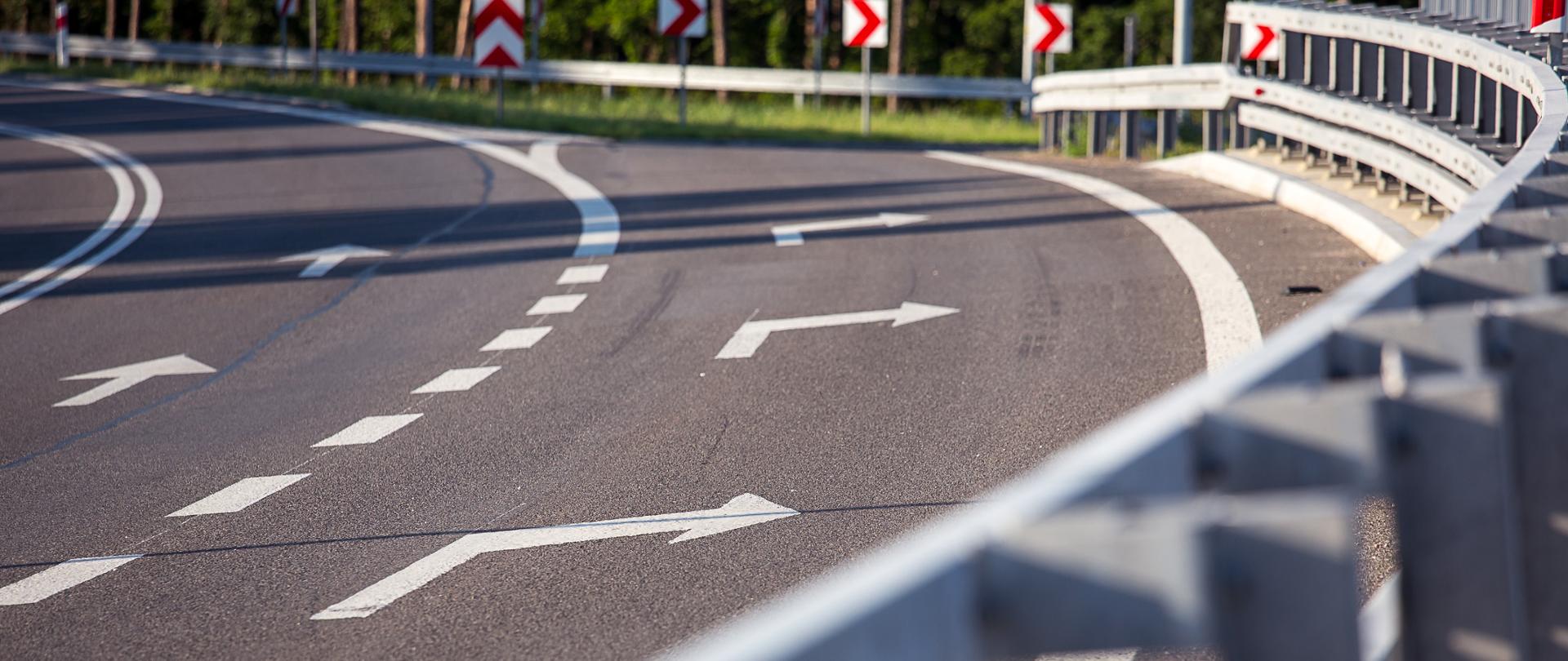 Zdjęcie z poziomu drogi. Pogoda słoneczna. Na pierwszym planie rozmyty widok na bariery energochłonne. Na drugim planie ostre, duże zbliżenie na oznakowanie na drodze – strzałki do skrętu w prawo. 