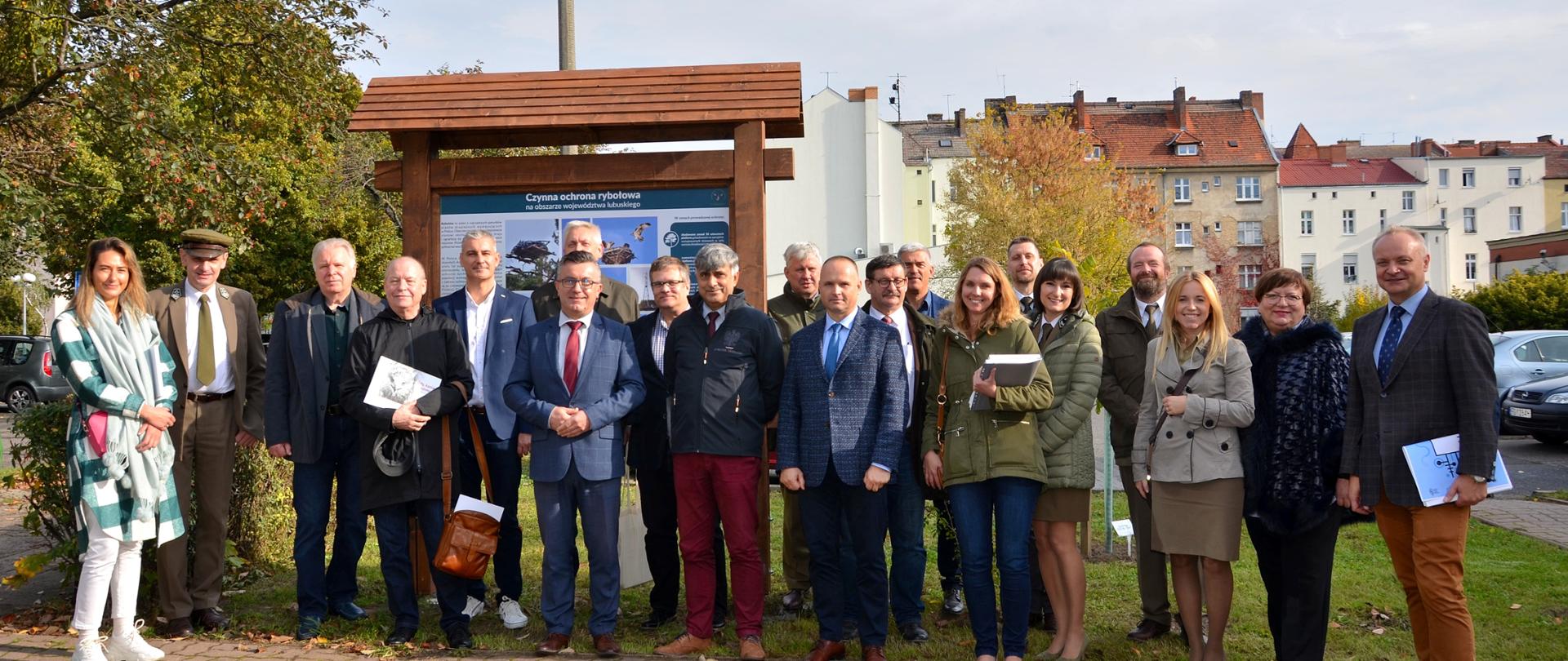 Grupa kilkunastu osób pozująca przed tablicą edukacyjną informującą o ochronie rybołowa. Wokół osób drzewa, w tle budynki.