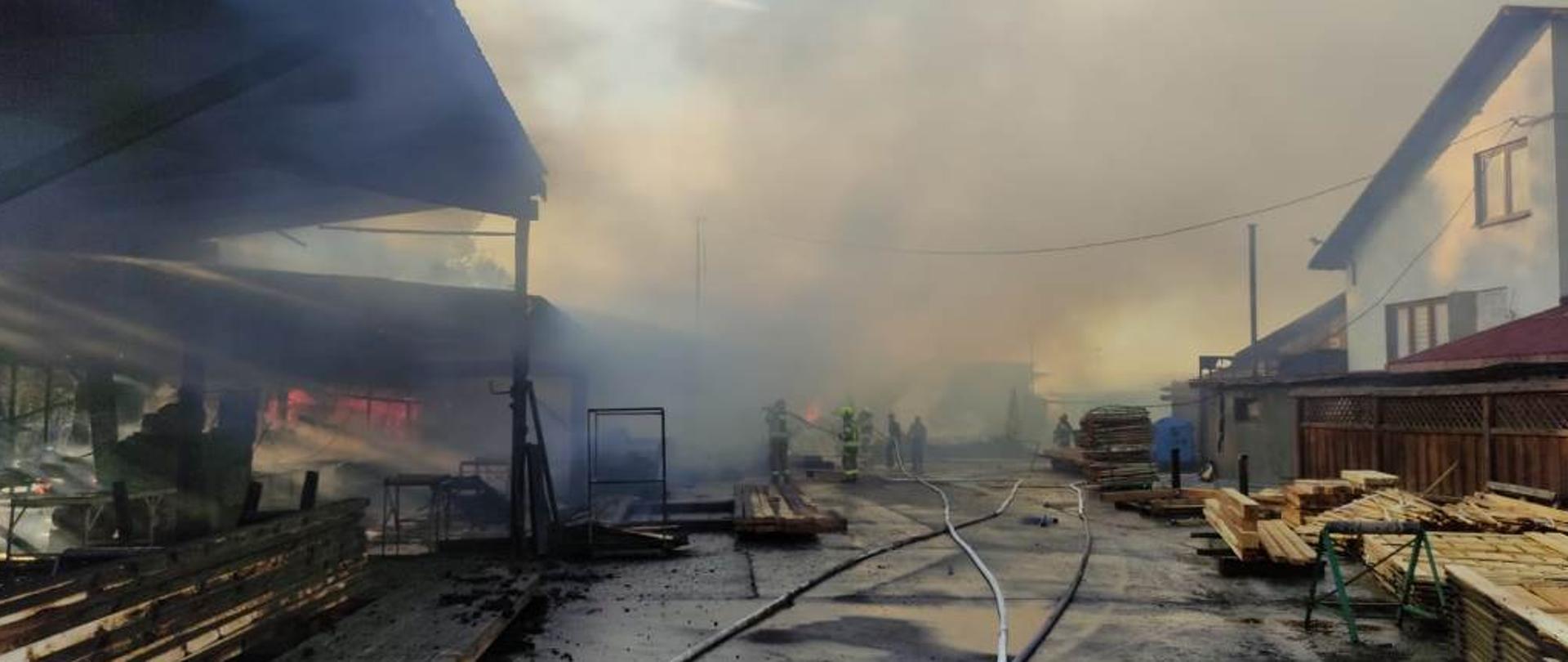 Teren tartaku, widoczne duże zadymienie, z prawej strony zdjęcia budynek mieszkalny, z lewej wiaty w których znajdowały się maszyny do przecierania drewna, na wprost zniszczona przez ogień hala oraz strażacy prowadzący działania.