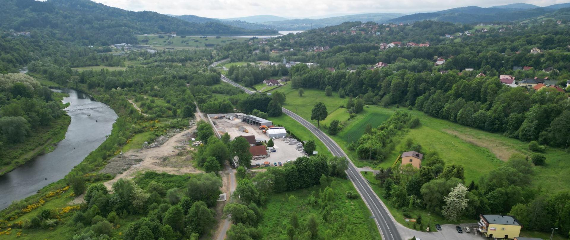 Widok z drona na drogę krajową 28 (odc. Wadowice-Sucha Beskidzka), widoczna zabudowa jednorodzinna zatopiona w zieleni oraz tereny leśne. W tle widoczne Beskidy. 