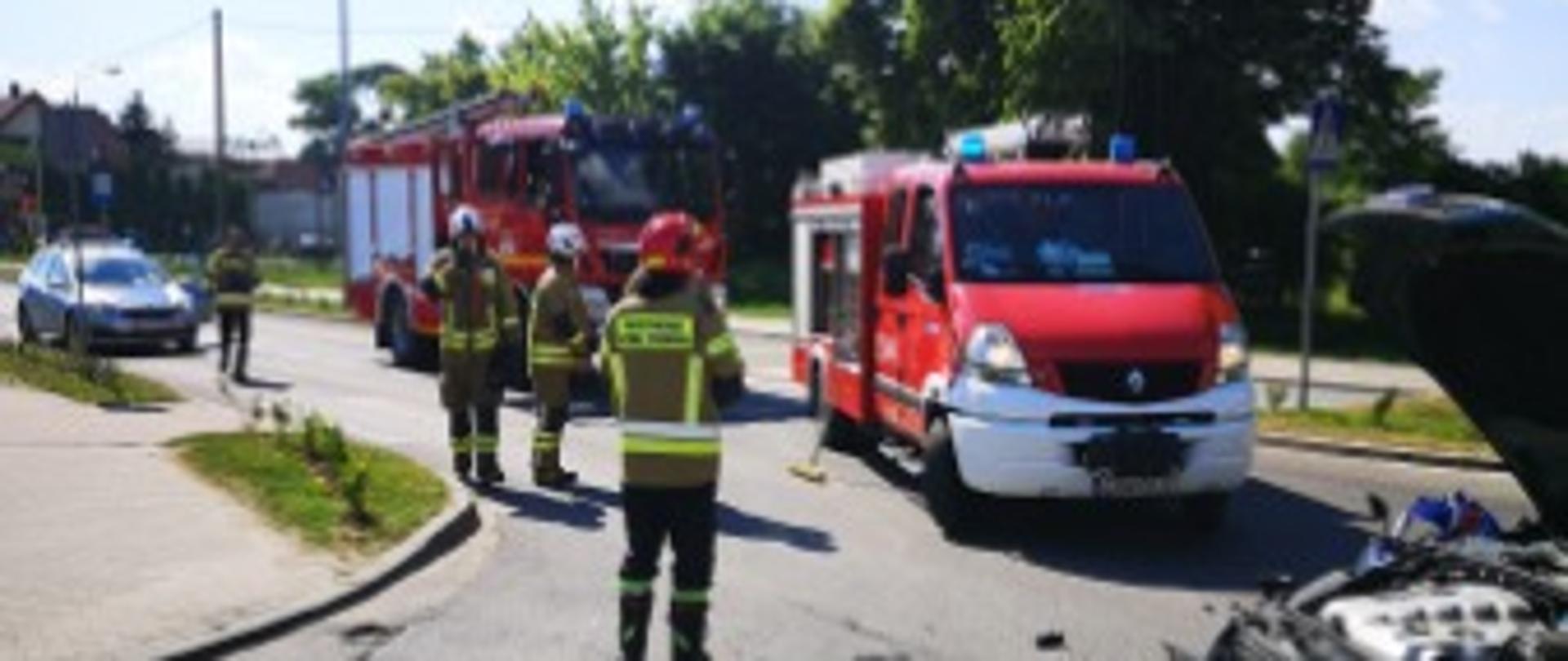 Zdjęcie przedstawia drogę asfaltową, na której stoją 4 samochody, 2 strażackie i 2 osobowe oraz 4 strażaków. Jeden z samochodów osobowych ma otwartą maskę.