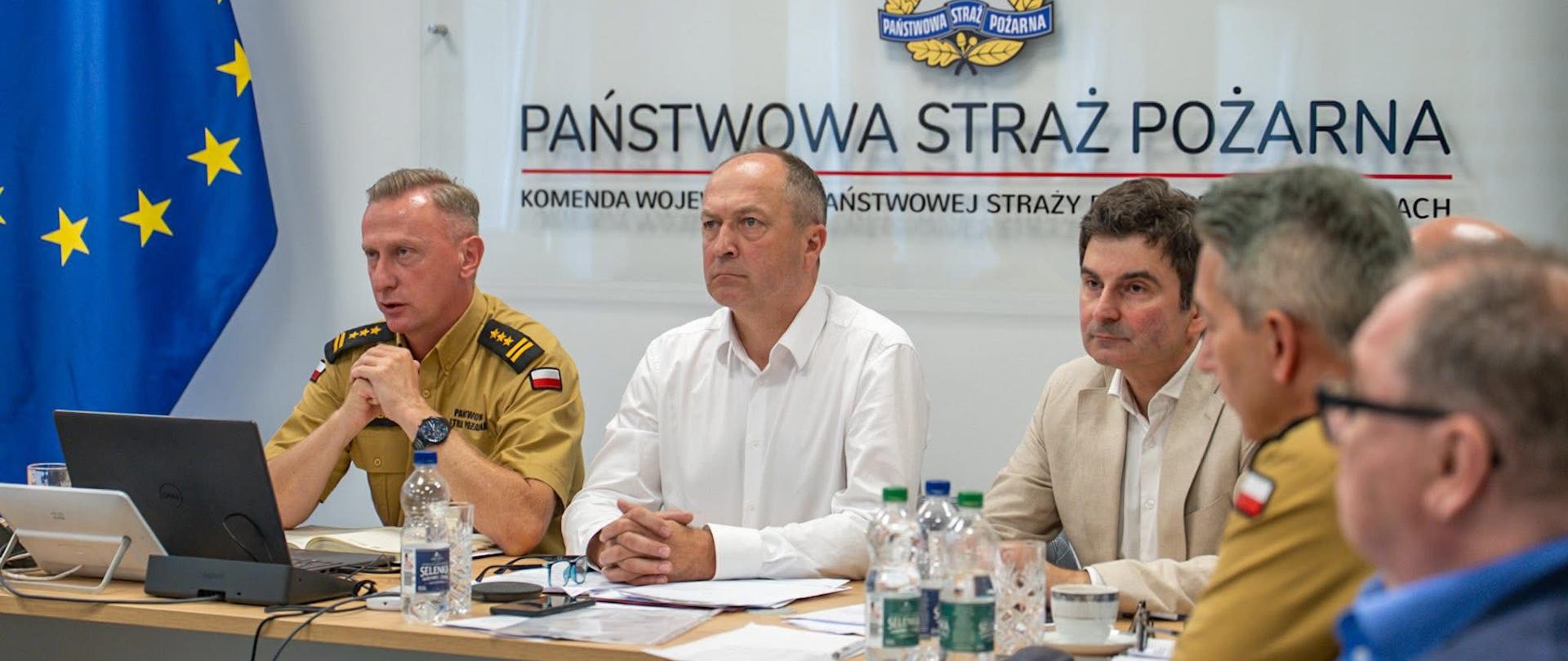 Na zdjęciu widać pięciu mężczyzn siedzących przy stole w sali sztabowej w Komendzie Wojewódzkiej Państwowej Straży Pożarnej w Katowicach. Trzech z nich jest wyraźnie widocznych na pierwszym planie, natomiast dwóch na drugim.Po lewej stronie, w beżowym mundurze służbowym, siedzi Śląski Komendant Wojewódzki PSP, Wiceprezes Zarządu Oddziału Wojewódzkiego ZOSP RP – st. bryg. Damian Legierski. Przed nim stoi laptop, butelka wody i telefon. W tle widać fragment flagi Unii Europejskiej z dwunastoma gwiazdami. Pośrodku siedzi Prezes Zarządu Oddziału Wojewódzkiego ZOSP RP – dh Henryk Kiepura w białej koszuli, który patrzy prosto przed siebie. Ma złączone dłonie na blacie stołu. W oddali, za nim, widać logo Państwowej Straży Pożarnej. Po prawej stronie, w jasnobeżowej marynarce, siedzi wojewoda śląski Marek Wójcik, który patrzy na osobę po swojej prawej stronie. Na stole przed nim stoi filiżanka, butelka wody i telefon.