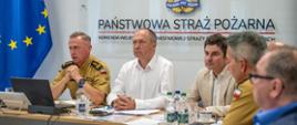 Na zdjęciu widać pięciu mężczyzn siedzących przy stole w sali sztabowej w Komendzie Wojewódzkiej Państwowej Straży Pożarnej w Katowicach. Trzech z nich jest wyraźnie widocznych na pierwszym planie, natomiast dwóch na drugim.Po lewej stronie, w beżowym mundurze służbowym, siedzi Śląski Komendant Wojewódzki PSP, Wiceprezes Zarządu Oddziału Wojewódzkiego ZOSP RP – st. bryg. Damian Legierski. Przed nim stoi laptop, butelka wody i telefon. W tle widać fragment flagi Unii Europejskiej z dwunastoma gwiazdami. Pośrodku siedzi Prezes Zarządu Oddziału Wojewódzkiego ZOSP RP – dh Henryk Kiepura w białej koszuli, który patrzy prosto przed siebie. Ma złączone dłonie na blacie stołu. W oddali, za nim, widać logo Państwowej Straży Pożarnej. Po prawej stronie, w jasnobeżowej marynarce, siedzi wojewoda śląski Marek Wójcik, który patrzy na osobę po swojej prawej stronie. Na stole przed nim stoi filiżanka, butelka wody i telefon.