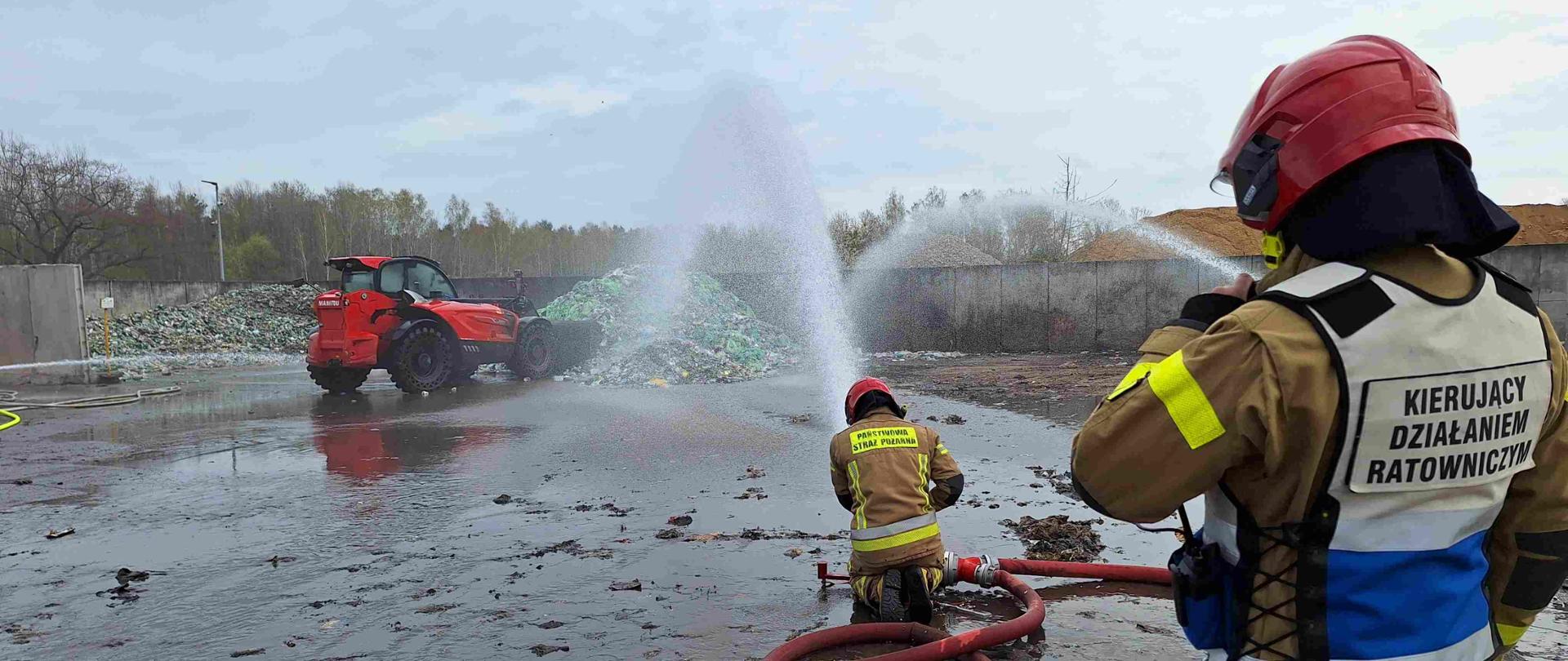 Strażacy ubrani w ubranie specjalne koloru piaskowego prowadzą działania gaśnicze palącej się hałdy odpadów.