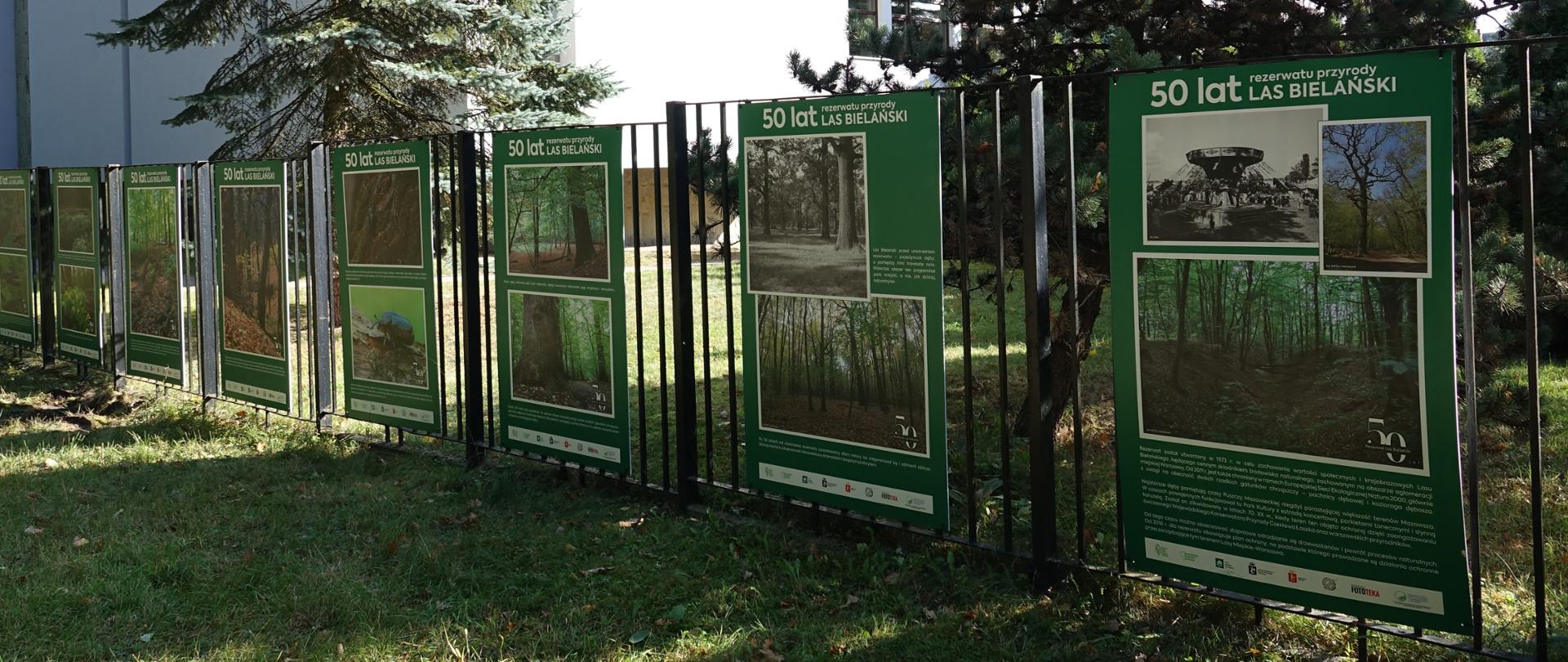 Plansze ze zdjęciami i tekstem zawieszone na ogrodzeniu, w tle budynek