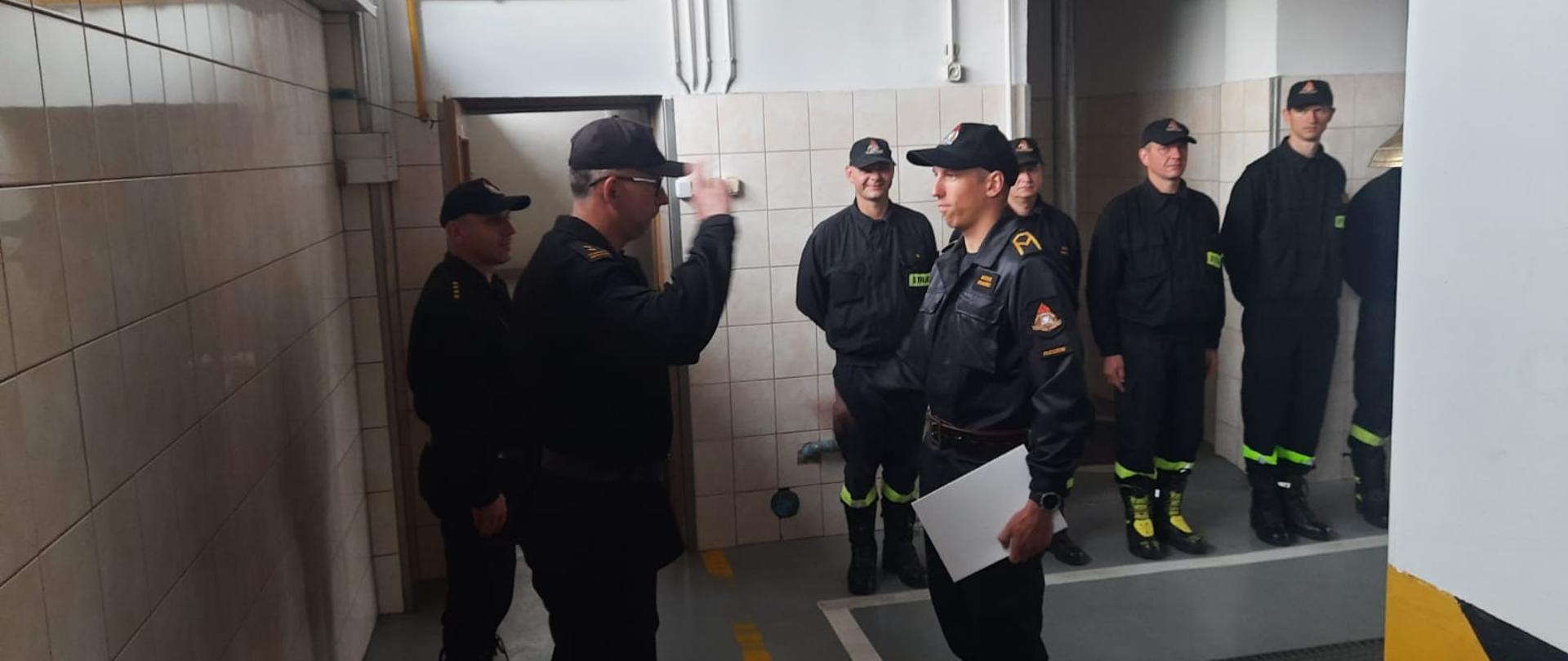 w centralnym planie stoi dwóch strażaków, z których stojący po lewej stronie salutuje strażakowi trzymającemu w ręku papierową teczkę na dokumenty, w tle stojący na baczność strażacy przyglądający sie ceremonii.