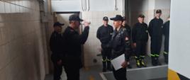 w centralnym planie stoi dwóch strażaków, z których stojący po lewej stronie salutuje strażakowi trzymającemu w ręku papierową teczkę na dokumenty, w tle stojący na baczność strażacy przyglądający sie ceremonii.