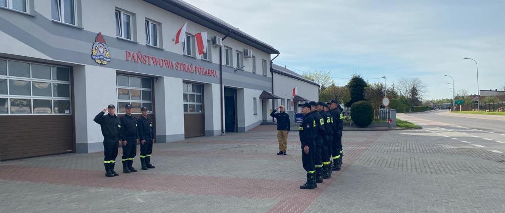 Zdjęcie przedstawia strażaków PSP przed budynkiem Komendy Powiatowej PSP w Lubaczowie. Strażacy stoją w zbiórce oraz oddają honor.