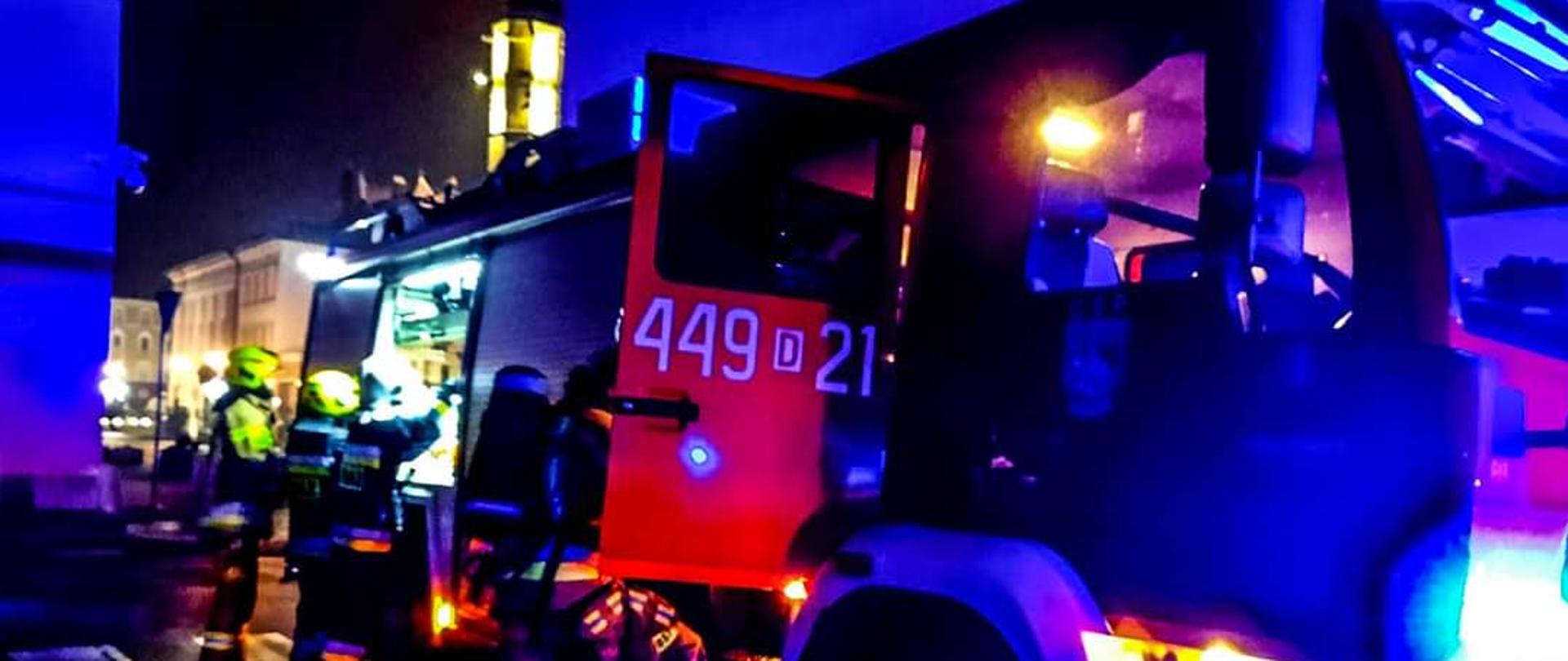 Obraz przedstawia samochód strażacki zaparkowany na ulicy. Pojazdy z włączonymi niebieskimi sygnałami alarmowymi. Przy pojeździe strażacy wyciągający sprzęt z bocznych skrytek. W tle widoczny jaworski rynek. Pora nocna.