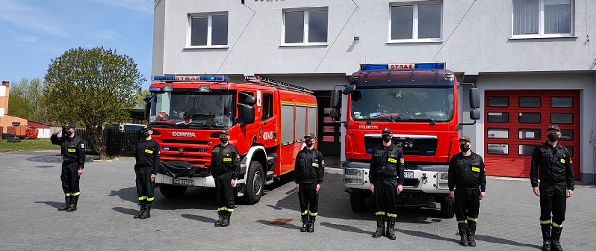 Zdjęcie przedstawia zmianę służbową przed obiektem strażnicy. W samochodach pożarniczych włączono sygnały świetlne. Strażacy stoją przed pojazdami w ubraniach koszarowych, na twarzach maseczki ochronne. Dowódca zmiany, pierwszy z lewej salutuje. Pomiędzy strażakami jest zachowany dystans społeczny.