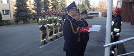 Podniesienie Flagi na maszt z okazji 103 rocznicy odzyskania niepodległości. Poczet flagowy w umundurowaniu galowym. W tle zbiórka strażaków w umundurowaniu bojowym.