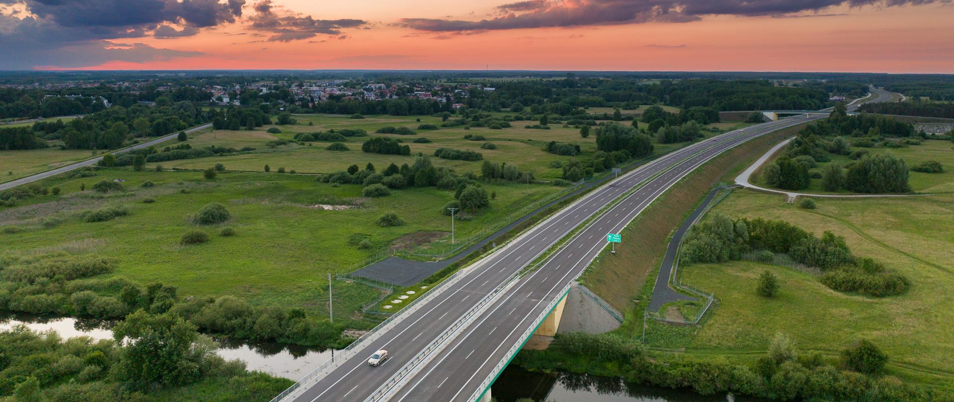Widok z lotu ptaka na drogę ekspresową S19 przecinającą zielone pola i rzekę; w tle zabudowania i zachód słońca z kolorowymi chmurami.