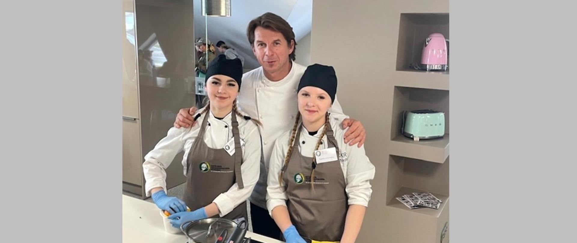 Dwie uczennice w strojach kucharskich pozują do zdjęcia z Karolem Okrasą.