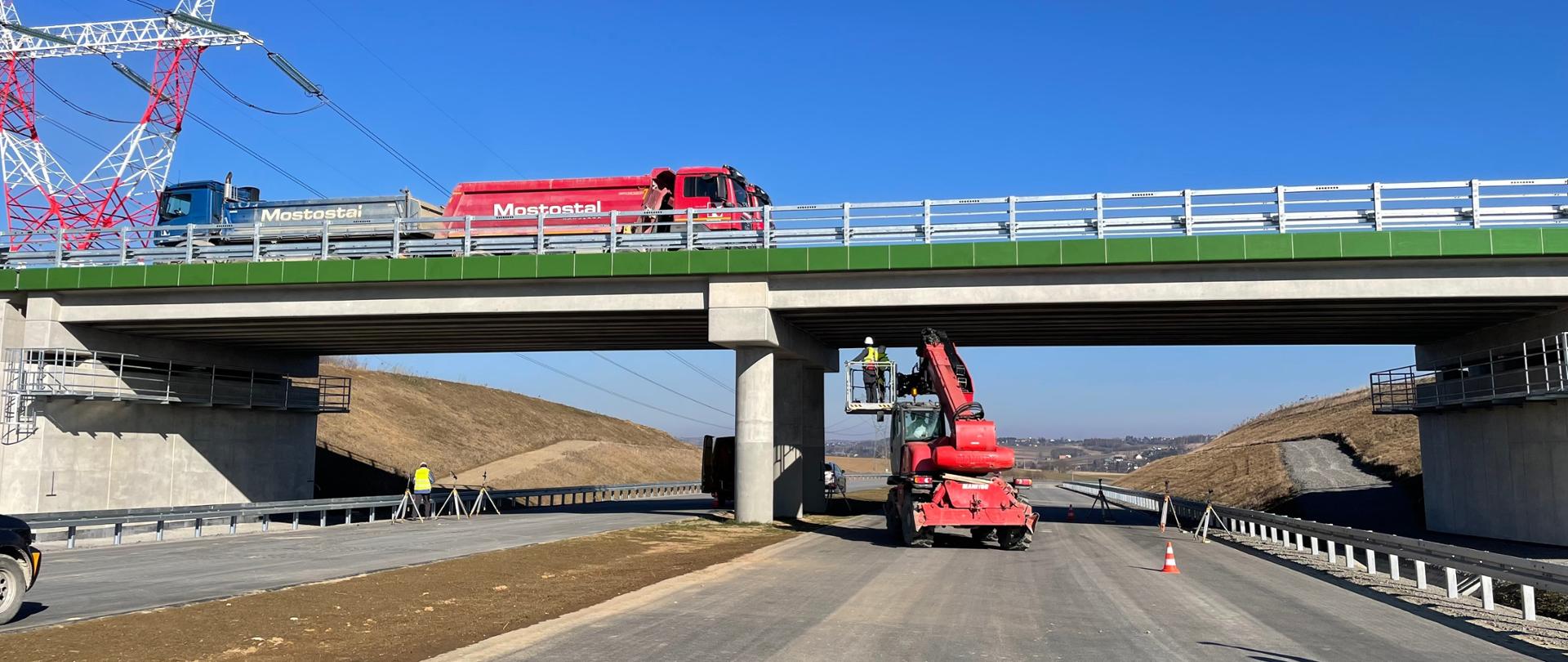 Zdjęcie wykonane na budowie drogi ekspresowej. Świeci słońce. Widok z poziomu ziemi na obiekt wymalowany na zielony kolor. Na wiadukcie stoją ciężarówki. W tym momencie trwa próbne obciążenie wiaduktu. Pod obiektem stoją ustawione sprzęty dokonujące pomiary oraz pracownicy. 