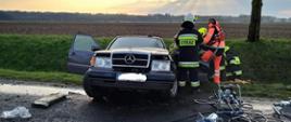 Na zdjęciu widoczny rozbity samochód osobowy marki Mercedes koloru szarego. Ratownicy wydobywają z niego poszkodowanego, który uprzednio został wycięty z pojazdu przy pomocy leżących z prawej strony narzędzi hydraulicznych.