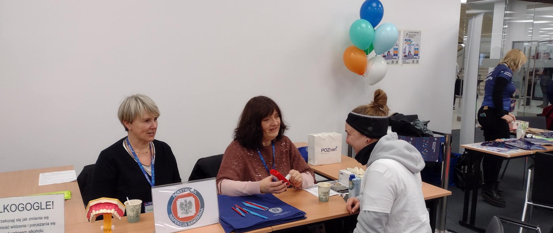 Za stołem siedzą 2 kobiety obsługujące stoisko, przed nimi kobieta korzystająca z oferty stoiska.
