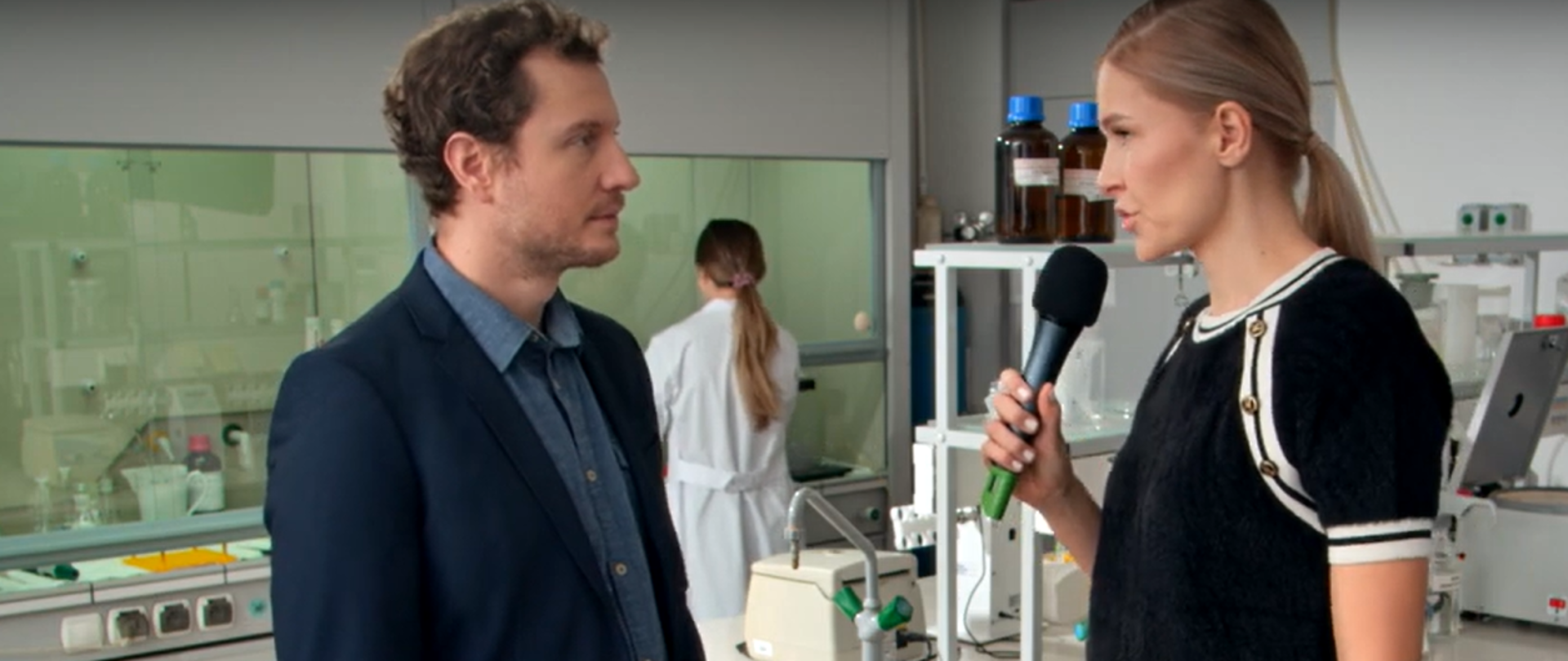 Fragment spotu. Dziennikarka zadaje pytania urzędnikowi, w tle sprzęt laboratoryjny i pracownica w fartuchu.