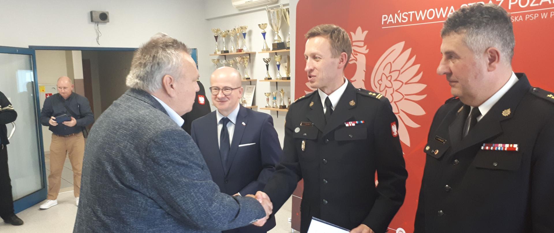 strażak ściska dłoń mężczyzny wręczając mu pismo, obok stoi drugi strażak w tle czerwony baner z orłem i pólka z pucharami