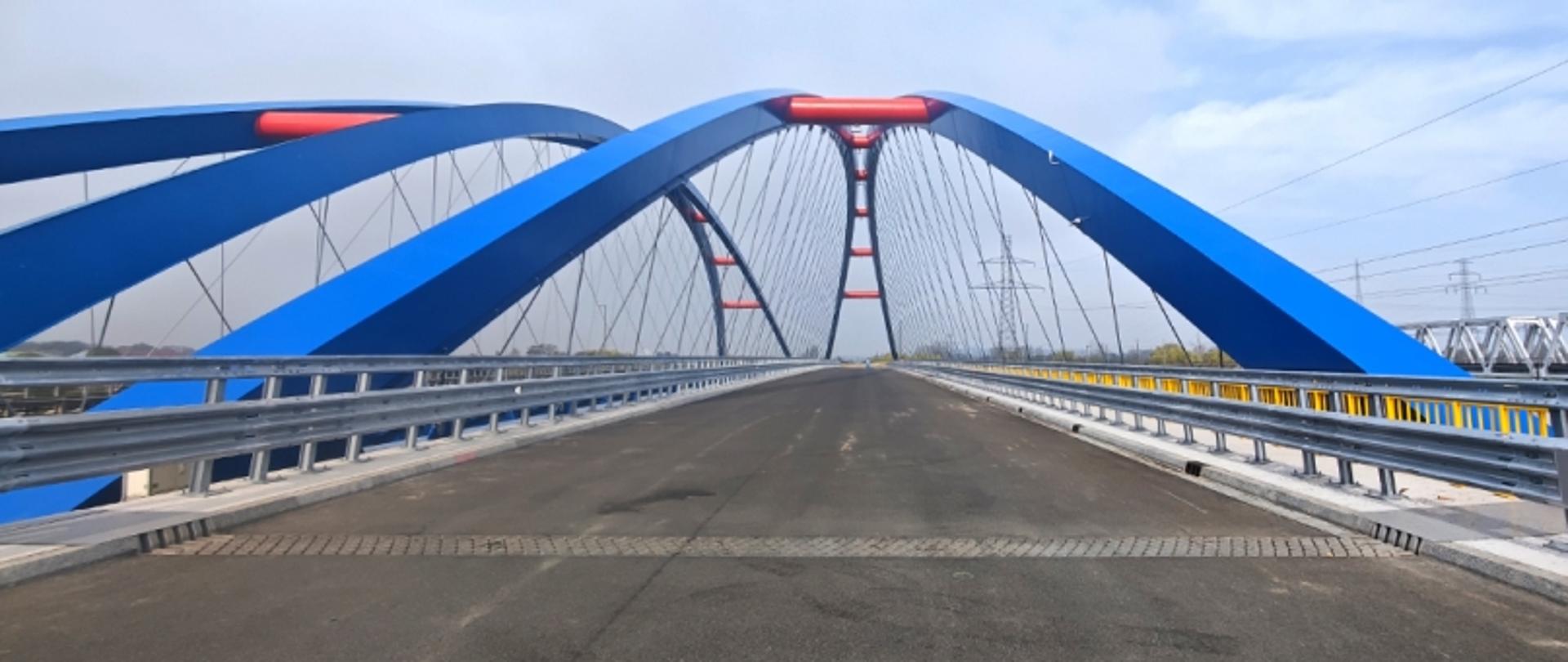 Pusta droga na moście z niebieskim elementem i metalowe barierki wyznaczające pasy jezdni.