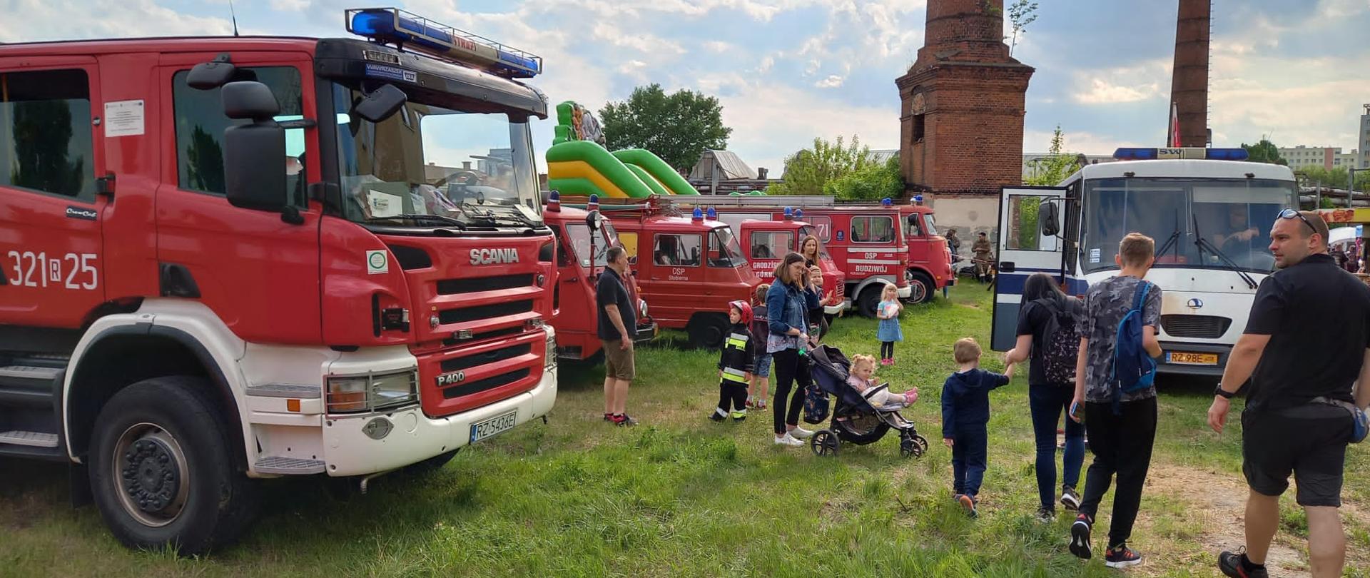 Zdjęcie przedstawia rodziny z dziećmi zwiedzające zabytkowe wozy pożarnicze i służby więziennej rozstawione na trawniku przed Muzeum Techniki i Militariów w Rzeszowie.