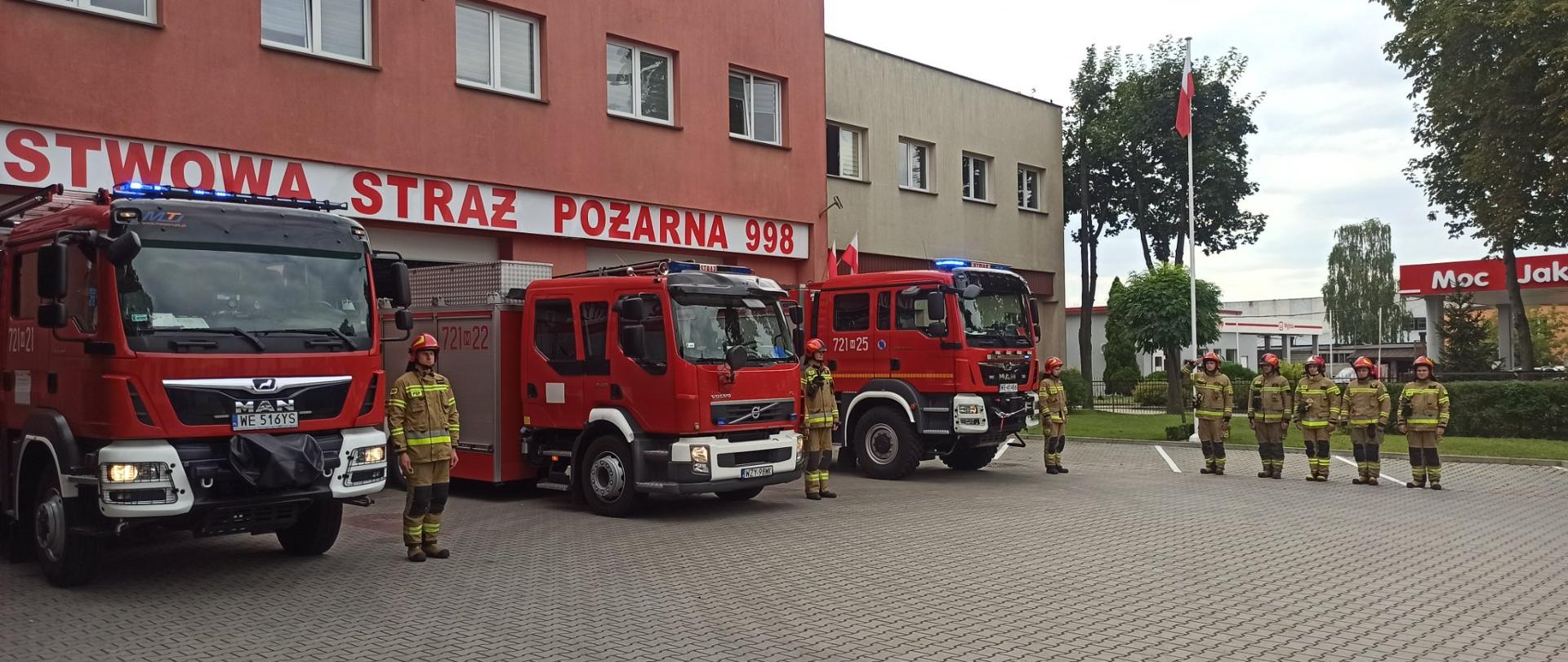 Trzy samochody bojowe stoją przed bramami garażowymi. Obok każdego z nich stoi jeden strażak. Pod masztem z wciągniętą flagą państwową stoją w rzędzie strażacy. Pierwszy z nich salutuje.