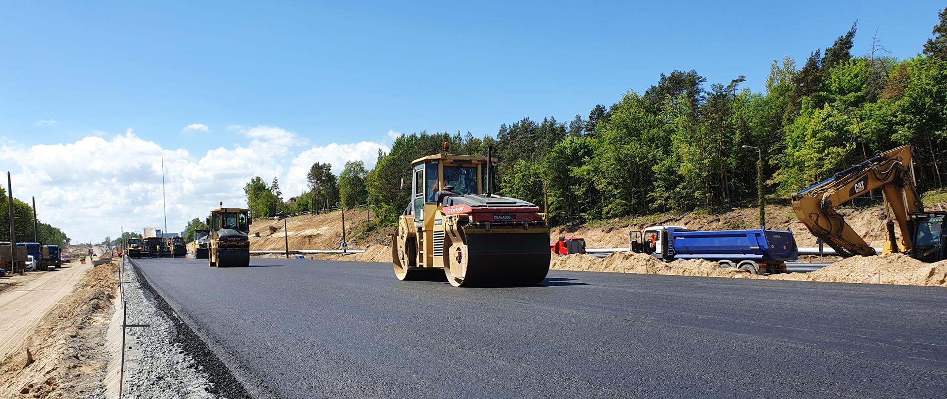 Na zdjęciu widać walce drogowe rozkładające asfalt na jezdni, z tyłu pracują koparki. Pogoda jest słoneczna.