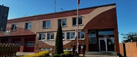 Na zewnątrz, przed budynkiem Państwowej Straży Pożarnej, po lewej stronie stoi w szeregu grupa strażaków w piaskowym umundurowaniu służbowym. W centrum, przy wysokim, biało-czerwonym maszcie, stoi trzech funkcjonariuszy w ciemnych mundurach wyjściowych, którzy salutują. Na maszcie znajduje się zawieszona flaga Polski. W tle widoczny jest dwupiętrowy, ceglasto-beżowy budynek straży pożarnej z czerwonymi bramami garażowymi oraz bezchmurne, jasnoniebieskie niebo. Na pierwszym planie po prawej stronie znajduje się chodnik z kostki brukowej oraz niska roślinność