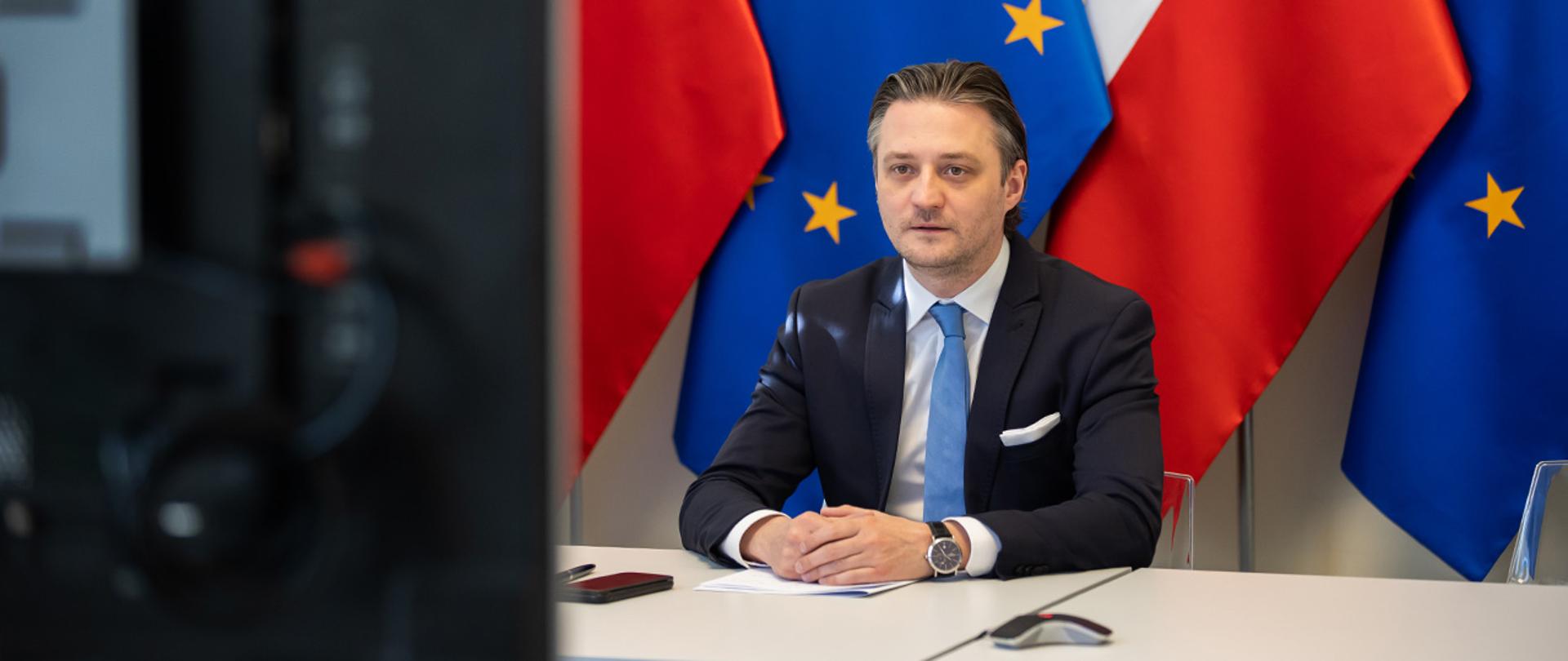 Na zdjęciu widać wiceministra Bartosza Grodeckiego siedzącego za stołem na tle flag Polski i UE.