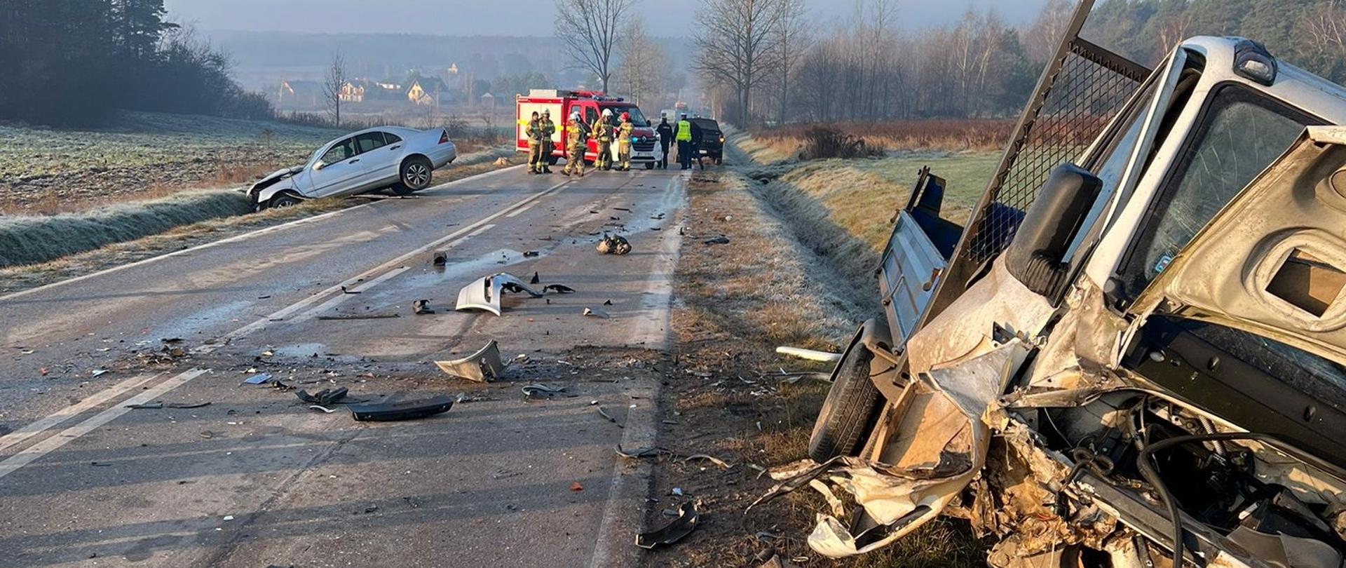 Zdjęcie przedstawia miejsce wypadku: po prawej stronie, na poboczu jeden ze zniszczonych samochodów, po lewej stronie, częściowo w rowie stoi drugie auto. Na jezdni rozrzucone fragmenty pojazdów, a w głębi stoją pojazdy ratownicze i ratownicy.
