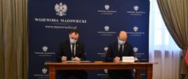 Zdjęcie przedstawia moment podpisania umowy. Za drewnianym stołem, na tle granatowej ścianki wizerunkowej Wojewody Mazowieckiego, siedzi dwóch mężczyzn ubranych w ciemne garnitury, jasne koszule i krawaty.