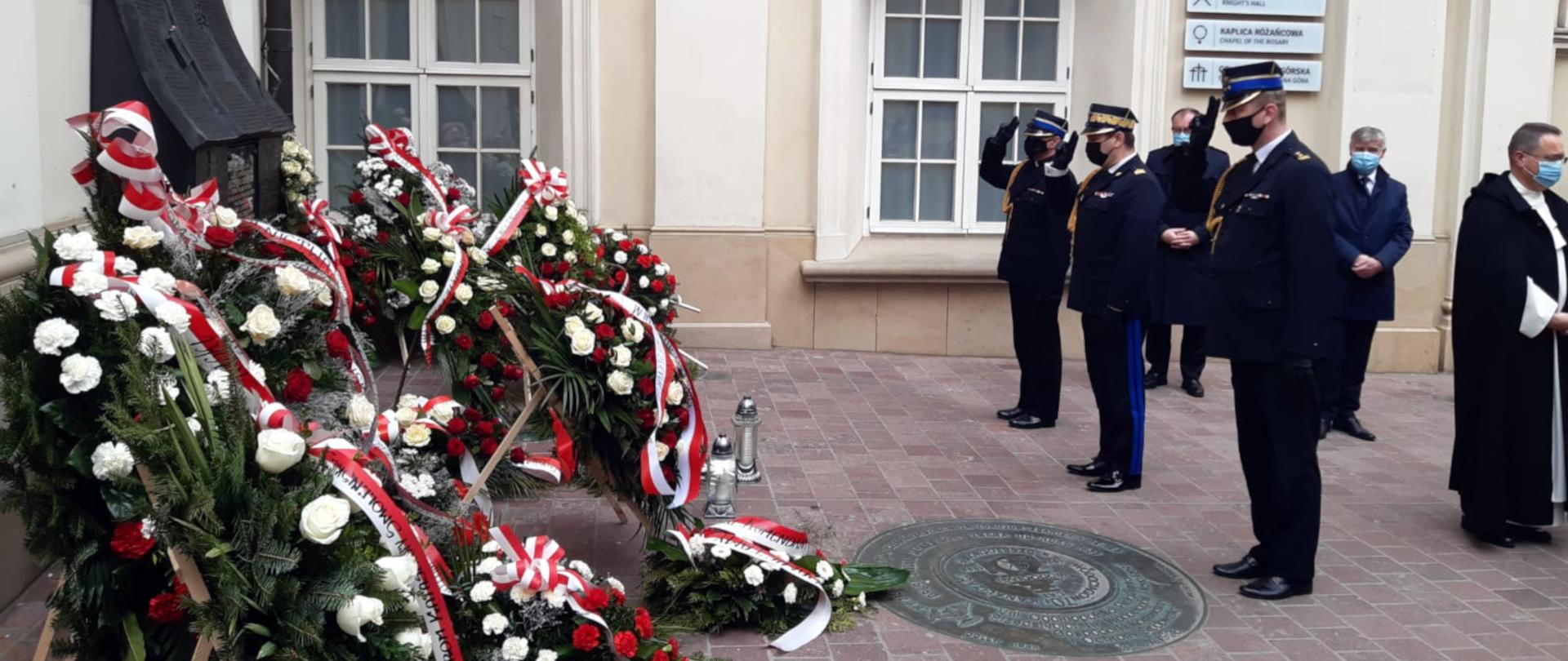 Na zdjęciu widoczny jest dziedziniec klasztoru na Jasnej Górze. Na dziedzińcu stoi w mundurach galowych delegacja PSP na czele ze śląskim komendantem wojewódzkim PSP oraz komendantem miejskim PSP i kapelanem krajowym strażaków wyznania ewangelicko- augsburskiego. Oficerowie salutuje przed tablicą upamiętniającą ofiary katastrofy smoleńskiej, przed którą złożone są wieńce i znicze.