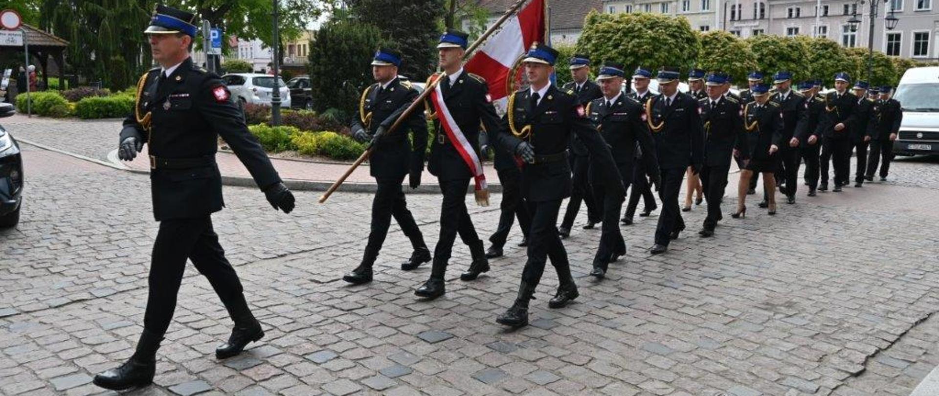 Strażacy w rzędzie maszerują przez miasto. Na przodzie idzie Dowódca Jednostki Ratowniczo-Gaśniczej. Strażacy mają ubrane mundury. Mundury są koloru czarnego. Strażacy mają ubrane czapki rogatywki. Czapki są koloru niebiesko-czarno-złotego. Za Dowódcą Jednostki Ratowniczo-Gaśniczej idzie trzyosobowy poczet sztandarowy. W środku pocztu sztandarowego funkcjonariusz ma na sobie biało-czerwoną szarfę. Funkcjonariusz w środku trzyma sztandar. Sztandar jest koloru biało-czerwonego. Strażacy znajdują się na tucholskim rynku. W tle widać budynki, drzewa, krzewy i samochody. Na ziemi znajduje się kostka brukowa. 