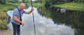 Mężczyzna stojący na drewnianym pomoście zanurza długą tyczkę w wodzie rzeki, otoczonej zielenią drzew i roślinności.