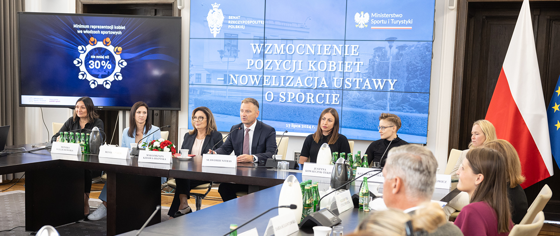 Zdjęcie z konferencji w Senacie: Wzmocnienie pozycji kobiet - nowelizacja ustawy o sporcie. Na pierwszym planie osoby siedzące przy stołach. W tle dwa duże monitory. W prawym górnym rogu flagi Polski i Unii Europejskiej.