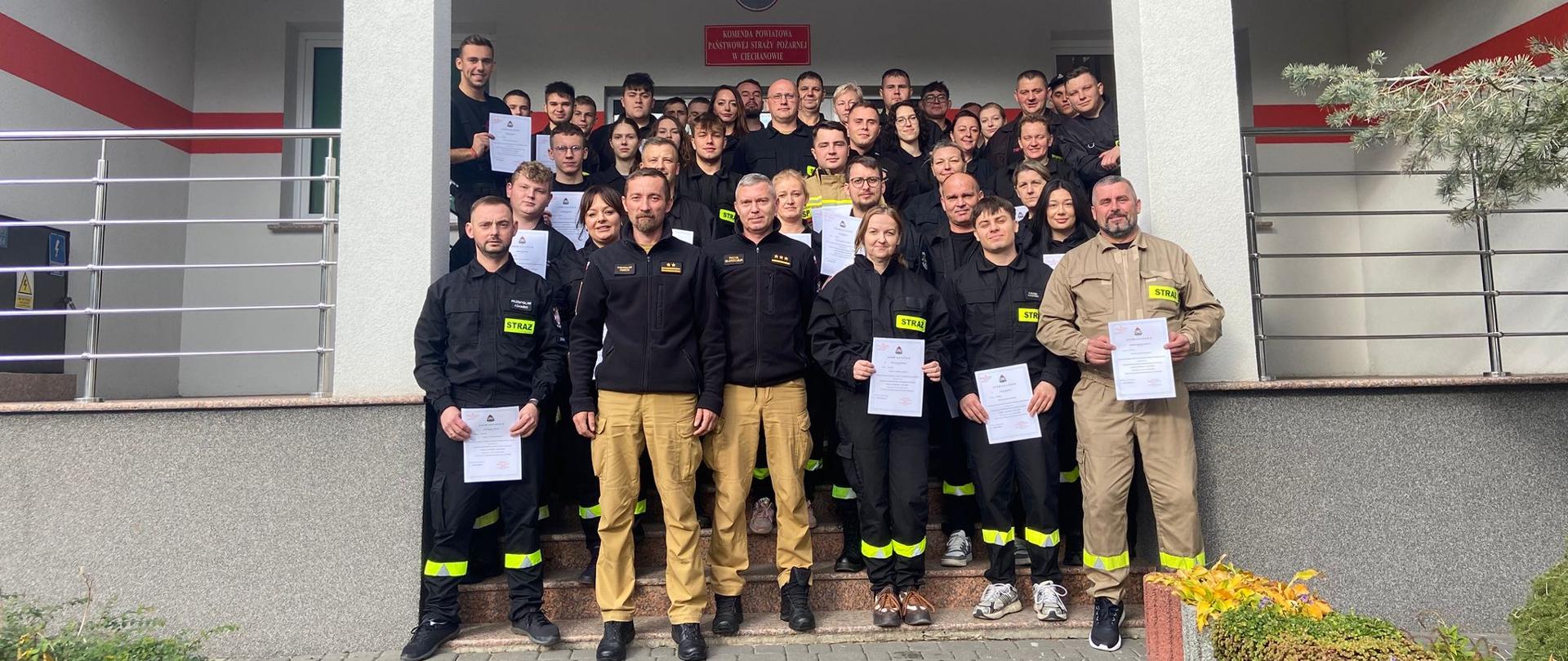 Nowi druhowie w strojach koszarowych przed wejściem do Komendy Powiatowej Państwowej Straży Pożarnej w Ciechanowie. Druhowie stojący jeden za drugim na schodach w grupie około 40 osób. na przedzie funkcjonariusze PSP 
