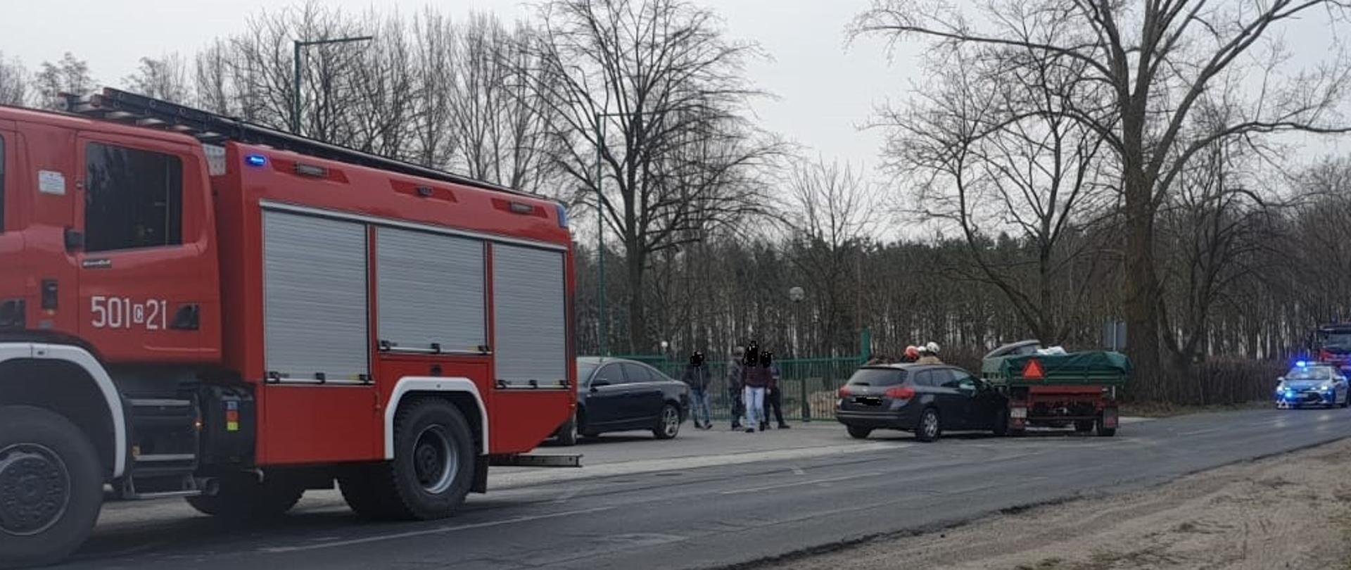 Zdjęcie na pierwszym planie przedstawia samochód osobowy oraz ciągnik rolniczy z przyczepą po kolizji drogowej. Na drugim planie radiowóz policyjny, dwa wozy straży pożarnej oraz funkcjonariusze policji oraz strażacy podczas czynności.