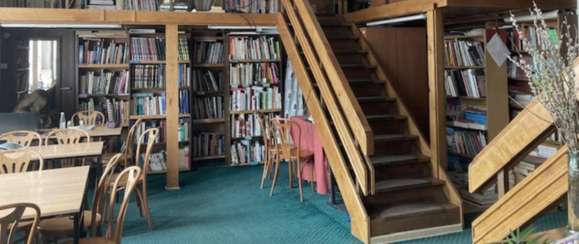 na zdjęciu jest widoczne wnętrze biblioteki szkolnej. Jest to dwupoziomowa sala z licznymi półkami na książki. Na antresolę prowadzą drewniane schody. Na podłodze znajduje się zielona wykładzina.