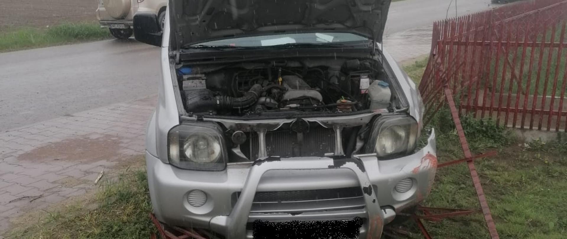 Na zdjęciu widać samochód osobowy, który uszkodził ogrodzenie posesji, ma on uszkodzony przód i otwartą maskę, w tle droga oraz przejeżdżające nią samochody.