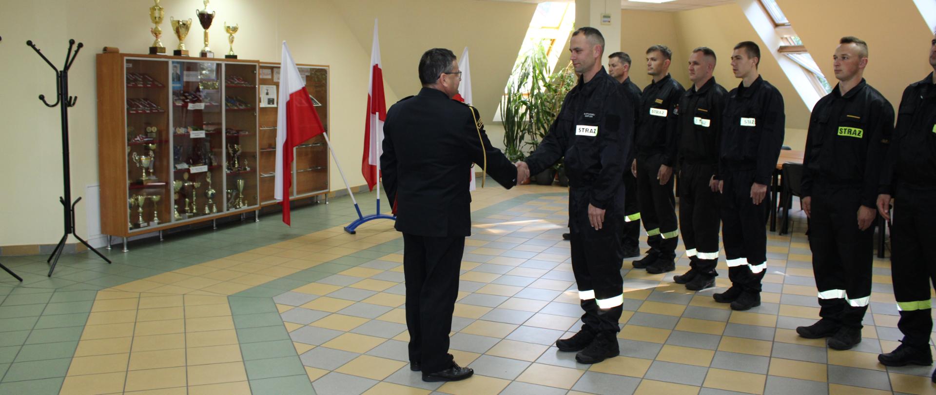 Komendant Powiatowy PSP w Golubiu-Dobrzyniu podaje dłoń strażakowi, który został przyjęty do służby. W tle widać ustawionych w szeregu uczestników zbiórki.