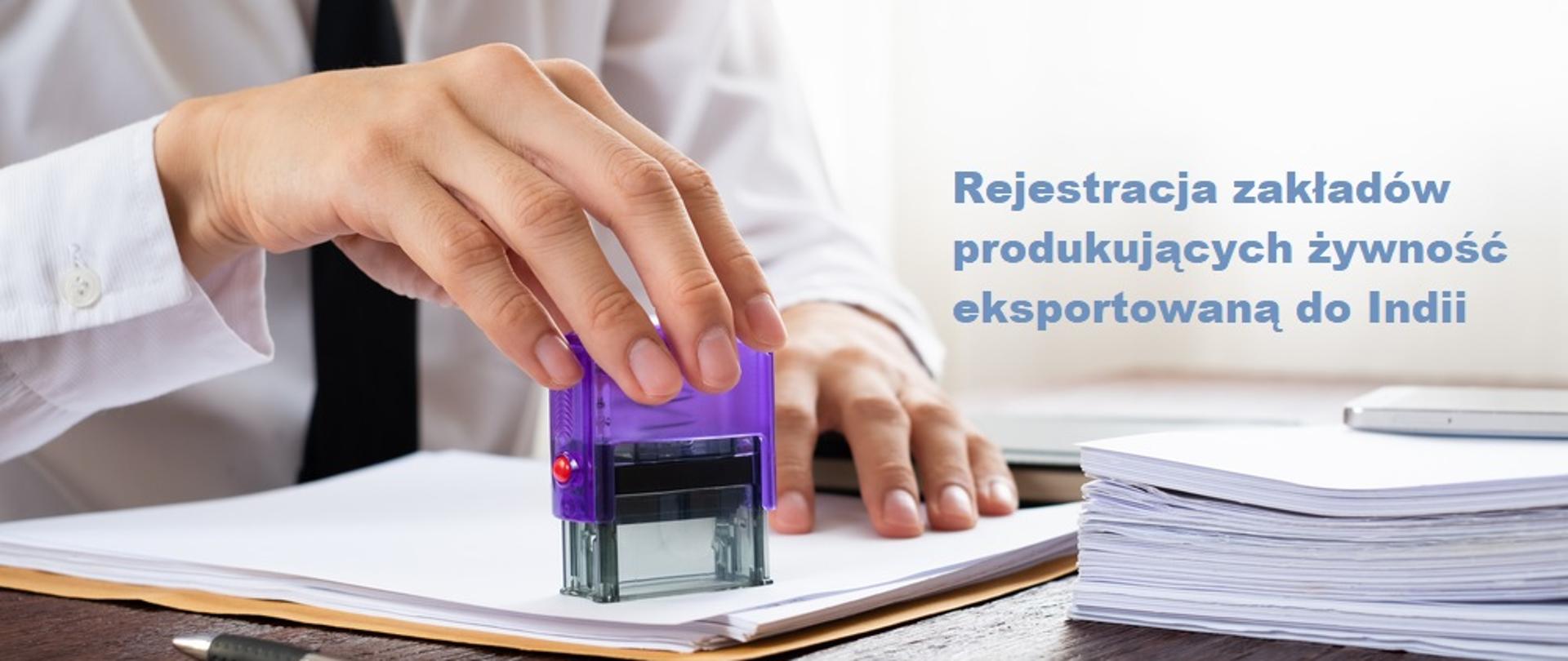 Na zdjęciu widać dłoń mężczyzny przybijającego pieczątkę na dokumencie leżącym na biurku, po lewej stronie leży również długopis, a po prawej inne dokumenty. Na zdjęciu widoczny jest napis: Rejestracja zakładów produkujących żywność eksportowaną do Indii