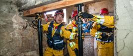 Strażacy Specjalistycznej Grupy Ratownictwa Technicznego "Brzeg" - trzej strażacy ubrani w żółte kombinezony z czerwonymi kaskami na głowie w trakcie stabilizacji części obiektu przy pomocy dwóch stempli budowlanych.